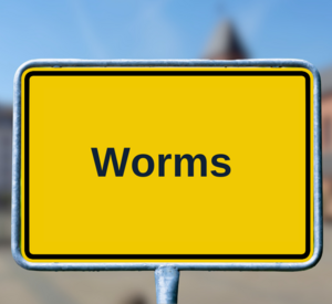 Zu sehen ist ein gelbes Straßenschild mit der Aufschrift Worms, im Hintergrund verschwommen zu sehen sind Häuser der Stadt Worms