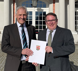Wolfgang Konder und ADD-Präsident Thomas Linnertz stehen nebeneinander vor dem Kurfürstlichen Palais in Trier. Sie halten mittig vor sich die Dienstjubiläumsurkunde von Wolfgang Konder.