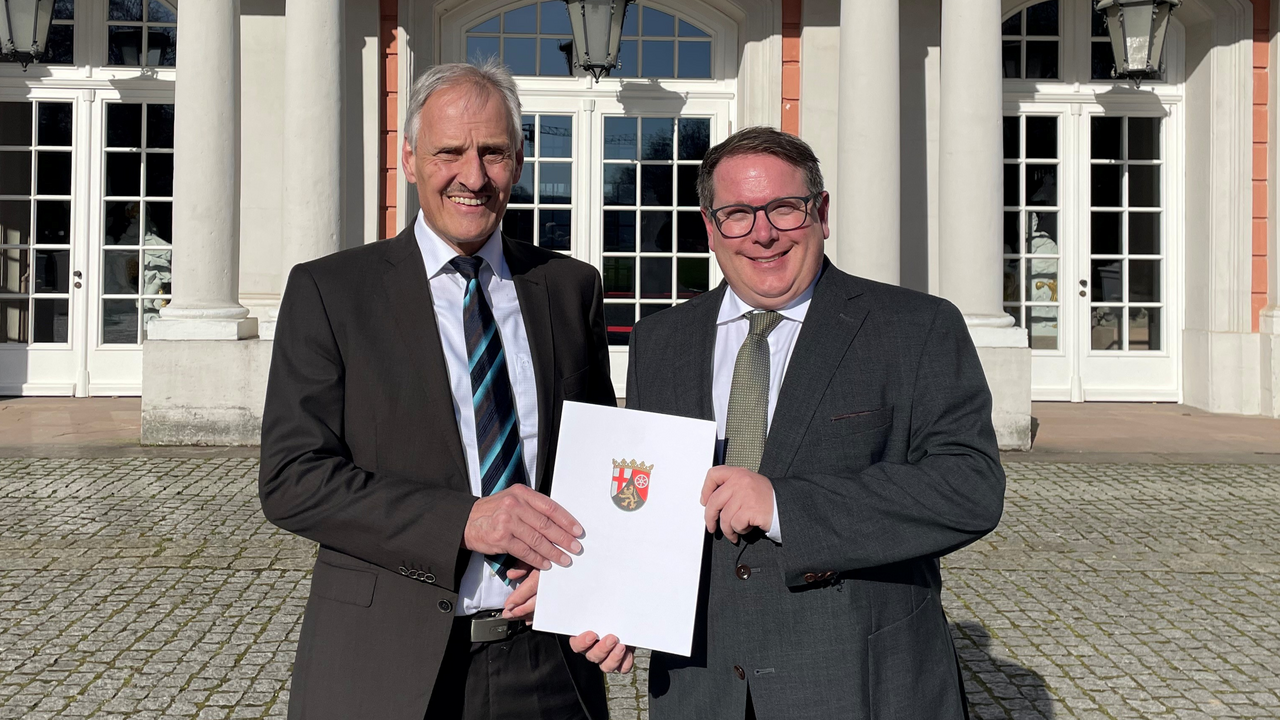Wolfgang Konder und ADD-Präsident Thomas Linnertz stehen nebeneinander vor dem Kurfürstlichen Palais in Trier. Sie halten mittig vor sich die Dienstjubiläumsurkunde von Wolfgang Konder.