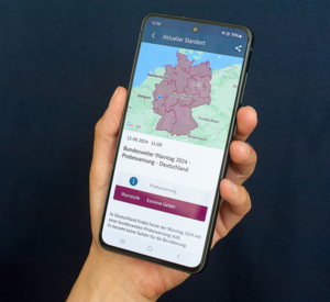 Symbolbild zum bundesweiten Warntag 2024 - ein Hand die ein Mobiltelefon festhält mit der Aufschrift bundesweiter Warntag 2024 und einer Karte