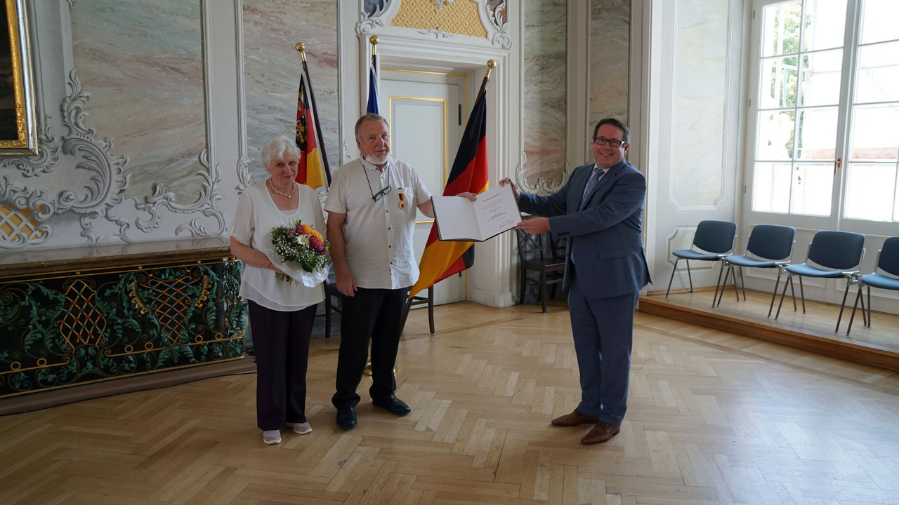 ADD-Präsident Thomas Linnertz übergibt Herrn Hans Muth die Urkunde und und Landesverdienstmedaille. Daneben steht die Ehefrau Renate Muth. Sie hat eine Blumenstrauß in der Hand.